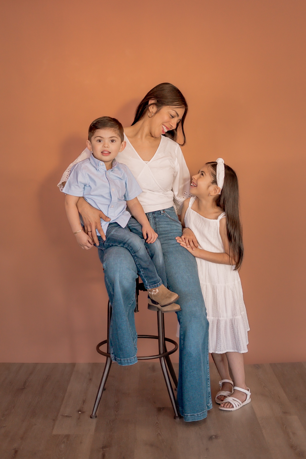 A mother in a white blouse and jeans seated on a stool with her young son and daughter against a warm terracotta studio backdrop.