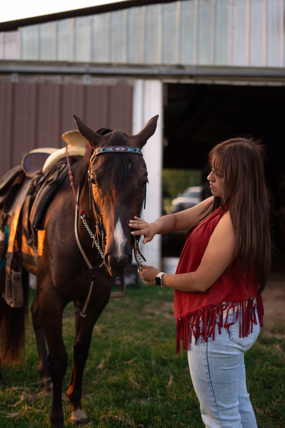 A woman in a red fringe top standing with a saddled horse at a barn in low golden light.